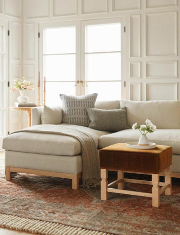 #showall::39840601768035 | The Ambleside cognac velvet ottoman sits atop a red Persian-inspired rug in front of a linen sectional sofa