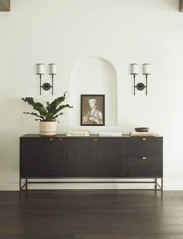 #color::black | The Rosamonde black wood sideboard with brown leather pulls and a metal base sits under an arched cut-out with a painting of a woman inside, flanked by two torchiere wall sconces.