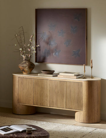 #color::natural-travertine | The Cagney sculptural vertical reeding media console styled with large scale artwork and a vase filled with botanicals.