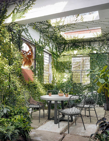 | The Agatha indoor and outdoor dining table with lavastone top and black iron base sits in a covered outdoor space with ivy-covered walls and black woven dining chairs