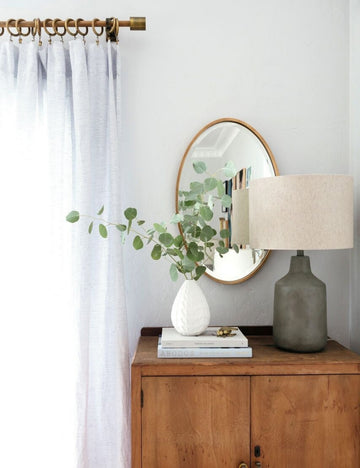| The Merci gold framed oval mirror is hung against a white wall behind a wooden side table and a white textured vase