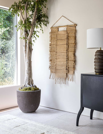 #color::natural #size::small | The Ukiah woven natural jute and hemp Wall Hanging hangs in a bright room with a ribbed lamp, a black wooden side table, and a plush white rug