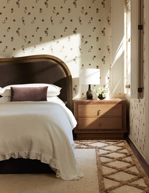 A wavy headboard bed frame and wooden nightstand sit in front of a wall covered with the Bamonte Textile String Floral Wallpaper by Jake Arnold
