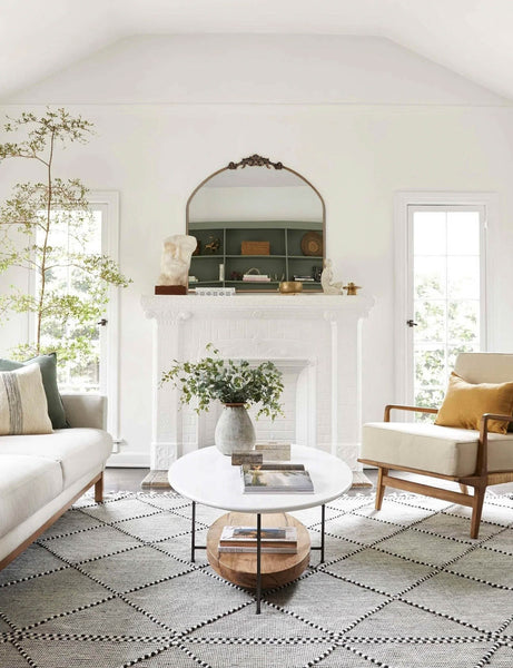 #color::gold | The Tulca arched gold mirror with flat bottom edge and traditional scroll detailing sits atop a fireplace in a bright living room with a natural sofa and accent chair and a black and white patterned rug.