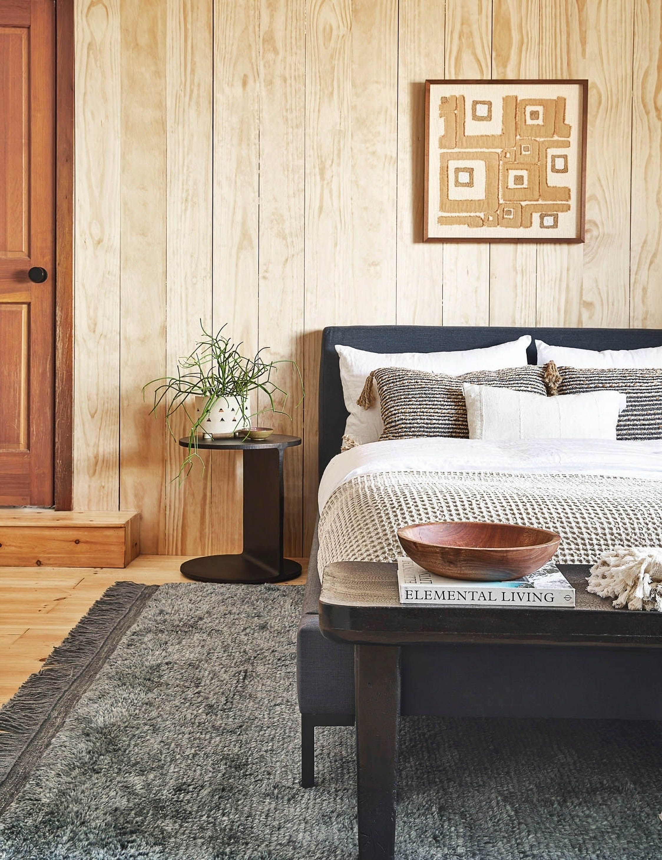 | The Illy metal geometric side table sits in a bedroom with a gray linen bed, a gray plush rug, and a light wood paneled wall