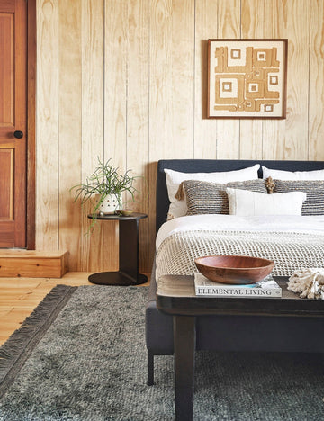 | The Illy metal geometric side table sits in a bedroom with a gray linen bed, a gray plush rug, and a light wood paneled wall