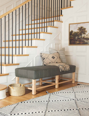 The ambleside loden gray linen bench sits by a staircase against white accented walls near a black and white patterned rug