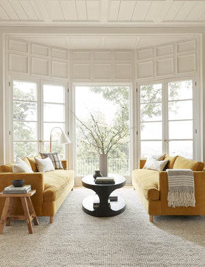 The Taos neutral light brown wool blend area rug lays in a white paneled living room under two gold velvet sofas and a black oval coffee table.