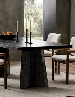 Hallen angular leg sculptural dining table styled with upholstered wood frame dining chairs, candlesticks and light colored rug.
