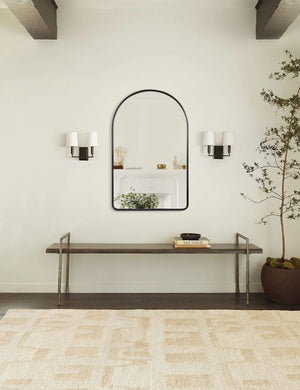 The Shashenka arched wall mirror with black frame hangs in a room with beamed ceilings above a metal bench