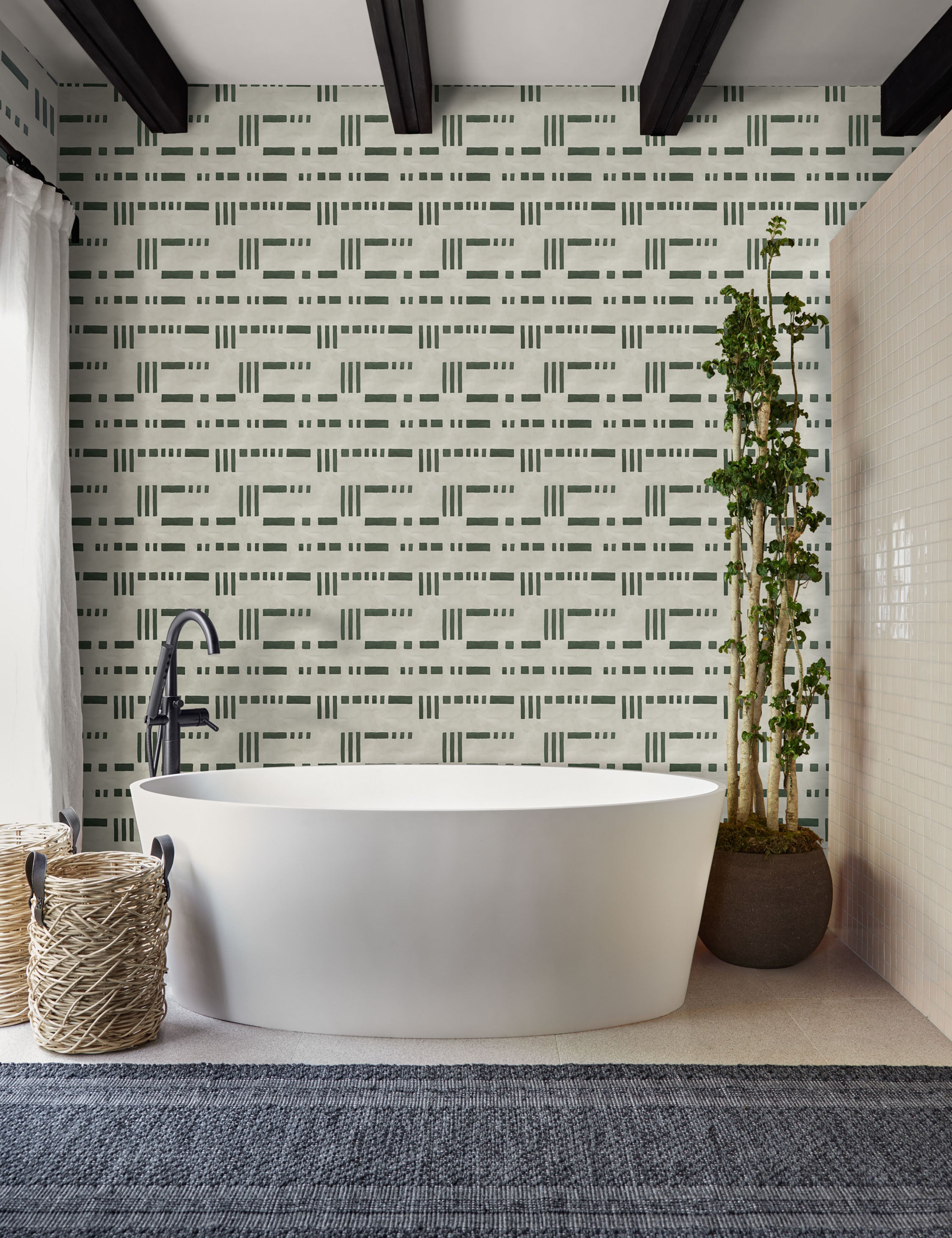 | The Artemis green and white dash-and-bar motif patterned wallpaper is in a bathroom with a white oval bathtub, a slate gray runner rug, and a natural tiled wall