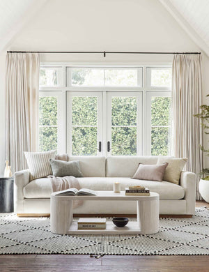 The Luna white-washed oak oval coffee table sits in a well-lit living room atop a white and black patterned rug in front of a white couch with long windows and french doors in the background.