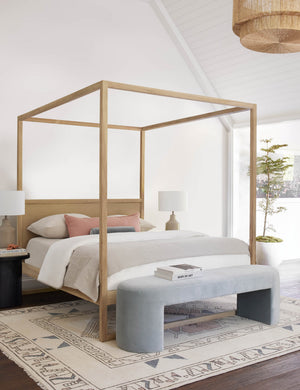 The Mikhail light blue velvet foam-padded bench sits in a bright bedroom with a light wood framed canopy bed atop a multicolored patterned rug.