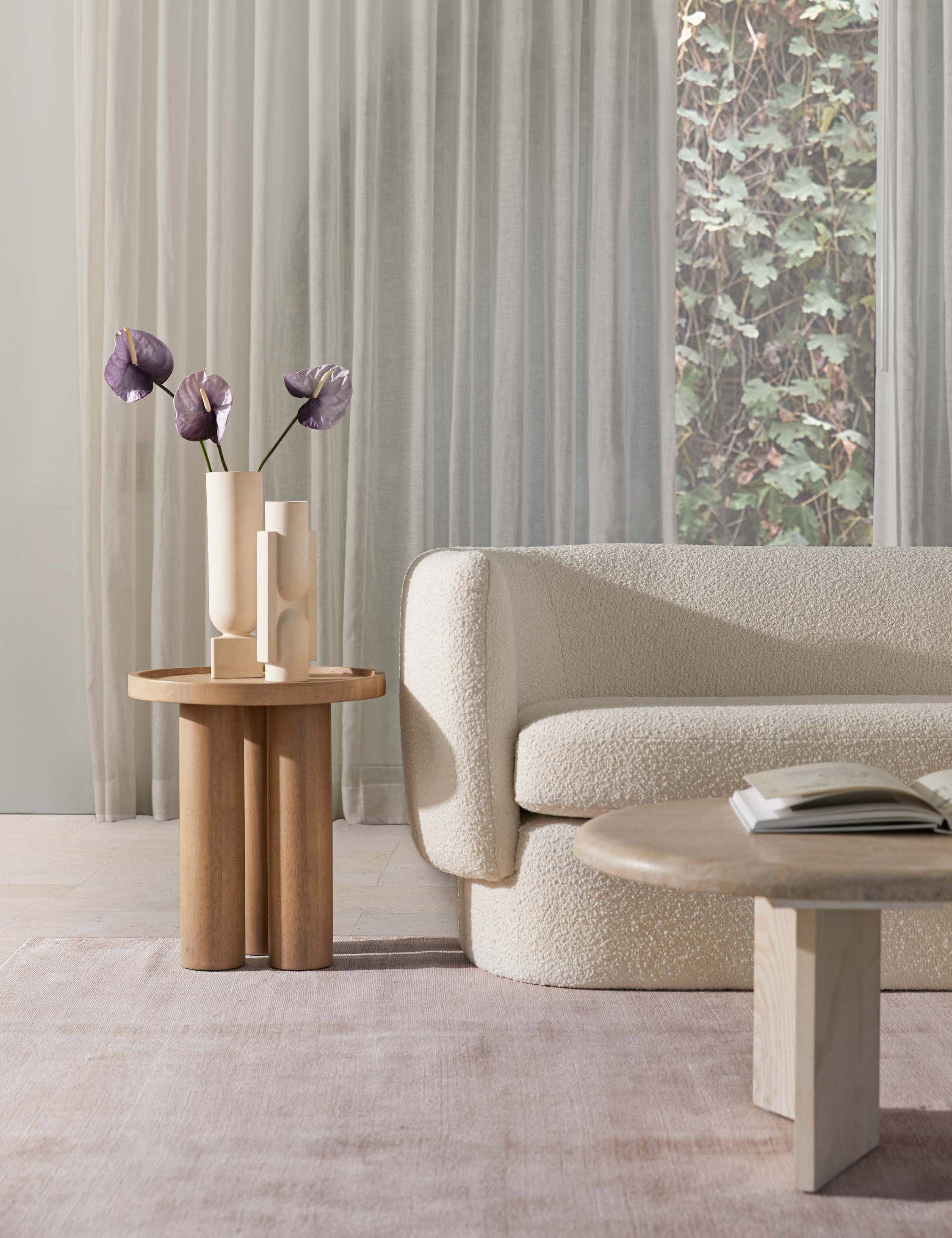 | The Delta natural wooden side table with pedestal base sits in a bright living room with a rounded white boucle couch, an ivory plush rug, and a whitewashed oval coffee table