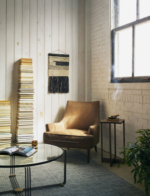 The Ilona Leather Accent Chair sits in the corner of a living room next to a round coffee table and two high stacks of books