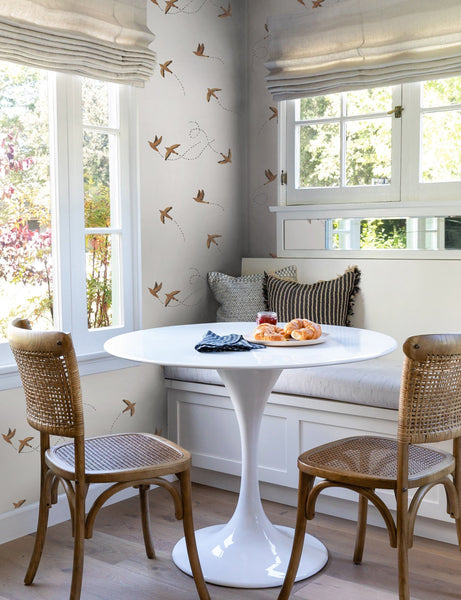 #color::rust | The Sparrow brown wallpaper by Rylee and Cru is in a dining nook with a white sculptural dining table, brown woven chairs that are surrounded by large colonial style windows.