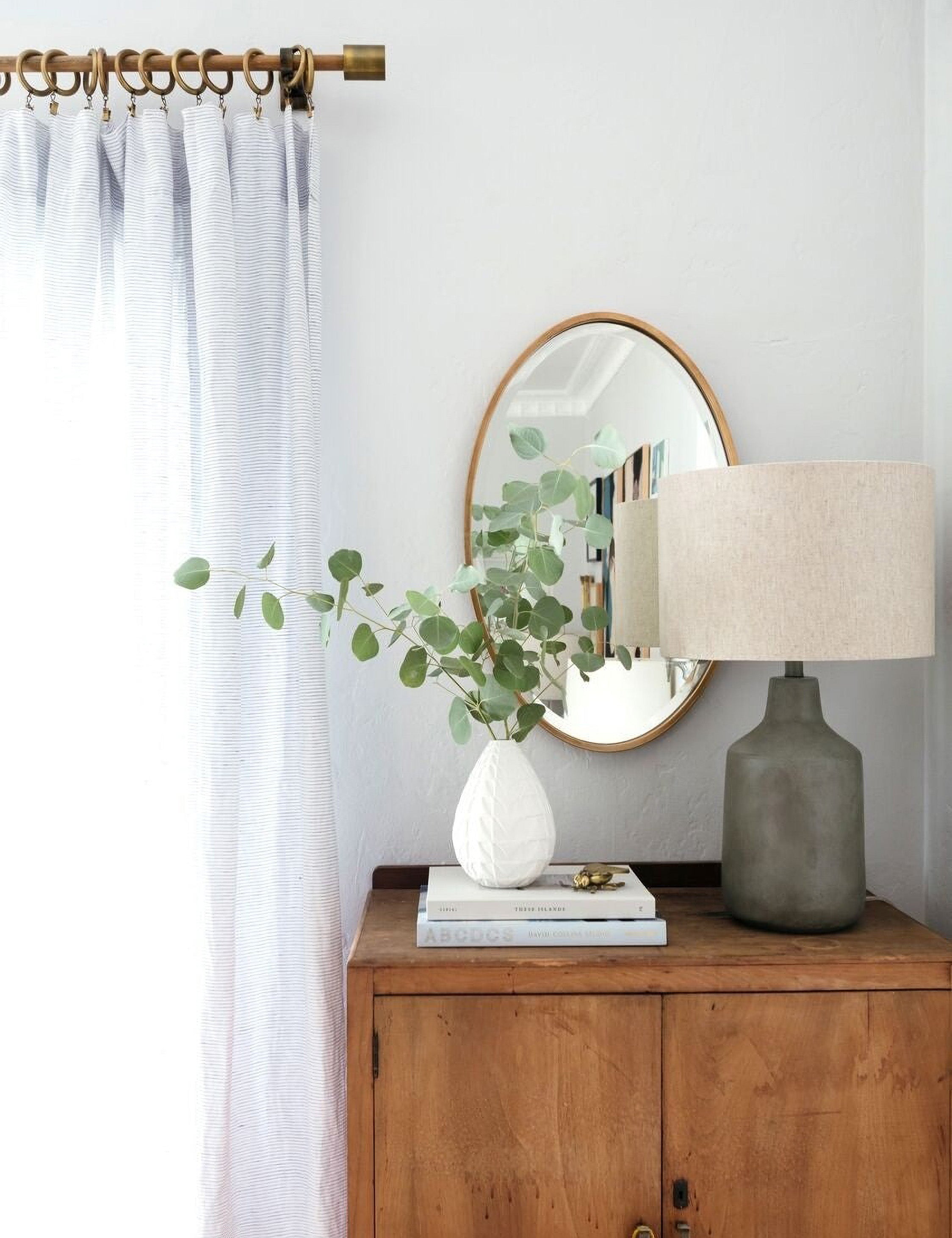 | The Merci gold framed oval mirror is hung against a white wall behind a wooden side table and a white textured vase