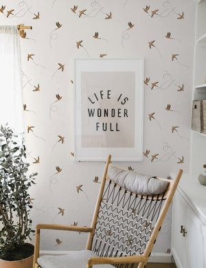 The Sparrow brown wallpaper by Rylee and Cru is in a room with a graphic wall art mounted against it behind a natural vase and a woven rocking chair with gray cushioning.
