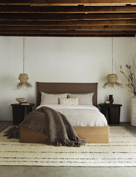 #showall::39490224390243 | The Nia mushroom brown linen bed sits atop a plush rug in between two jute pendant lights under a wooden beamed ceiling