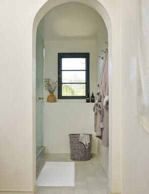 The Cloud loom sustainable bath mat by coyuchi lays on a white tiled floor in a bathroom with an arched entryway