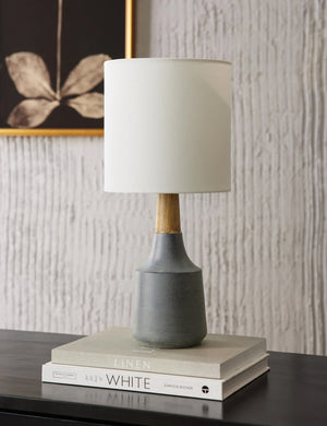 The Marcella gray vase-shaped Mini Table Lamp with ceramic base sits on top of a wooden sideboard with a stack of books and a floral wall art in the background