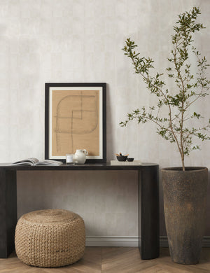  The Vela beige all-natural jute and cotton textural pouf sits in a room under a black wooden console table with a earthenware vase and a brown patterned wall art