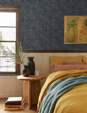 The Vadala long lumbar pink patterned pillow lays atop a bed with mustard-toned linens in a bedroom with blue patterned walls and a sculptural wooden nightstand