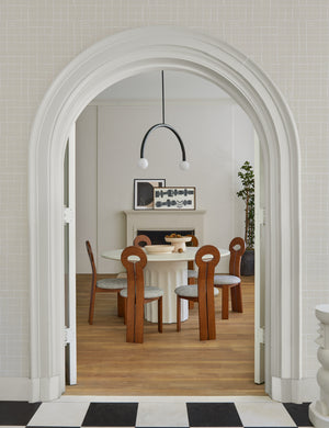 The Whit honey wood sculptural dining chair by sarah sherman samuel sits in a dining room surrounding a white circular dining table with a white fireplace in the background.