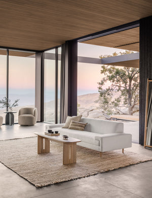  The Estee white linen upholstered sofa sits atop a jute rug in a room with wrap around floor to ceiling windows 