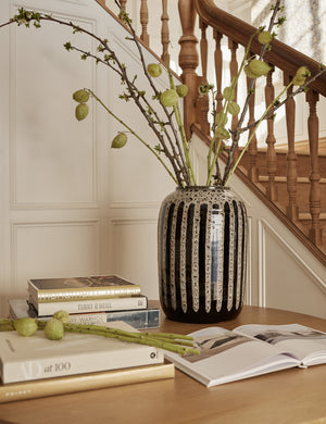 Wheaton full-bodied black and natural striped vase with a plant inside sits on a table with stacks of books and a staircase in the background