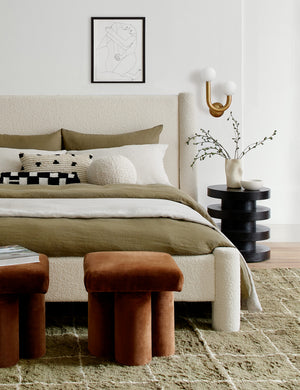  Ivory Bouclé Ball Pillow by Sarah Sherman Samuel sits on a natural linen framed bed in a bedroom with a moss green rug and conganc ottomans