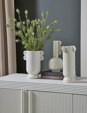 The Marguerite matte white ceramic vase with a grooved neck sits with other white vases atop a textured white sideboard