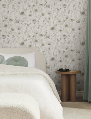 The Scalamandre Lo soft and pastoral gray toned floral wallpaper is in a bedroom with a natural linen framed bed, natural and cream toned bedding, and a wooden pedestal-base nightstand