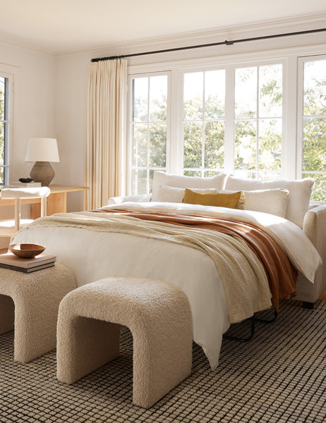 #color::cream | Two Tate Cream Boucle stools sit at the end of a bed on a gridded rug with a wooden bowl and a stack of books on one of them