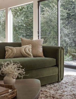 The Kisha natural-toned square throw pillow sits on a green velvet sofa with a plush ivory throw pillow