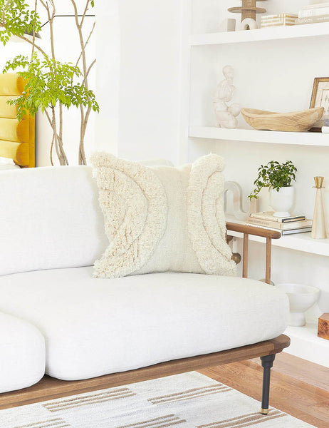 #color::natural #size::square | The arches ivory square pillow sits atop a white linen sofa with a wooden frame with a decor filled shelf in the background