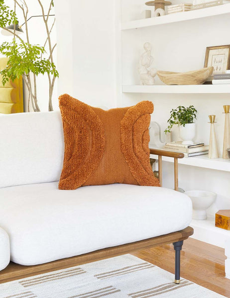 #color::rust #size::square | The arches rust square pillow sits atop a white linen sofa with a wooden frame with a decor filled shelf in the background