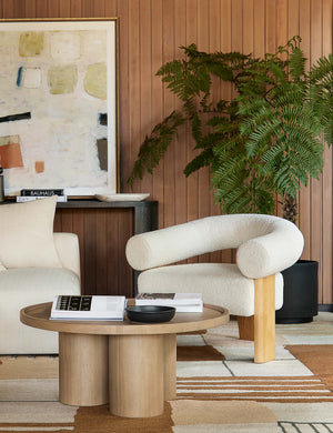 The Delta ash wood round coffee table with three column legs sits in a living room with wood paneled walls, a brown geometric patterned rug and a rounded sherpa accent chair.