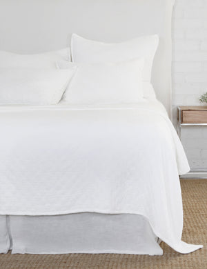 The Ojaj white cotton matelassé coverlet by pom pom at home lays atop a white linen framed bed in a bedroom with a jute area rug, a white brick wall, and a wooden nightstand