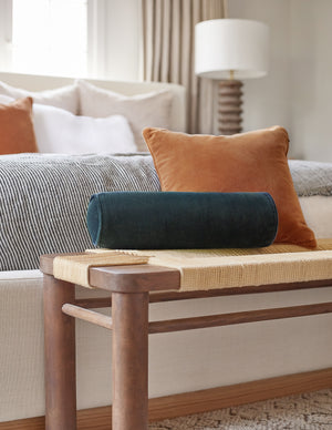 The Sabine navy velvet cylindrical bolster pillow sits atop a woven bench with a burnt orange velvet throw pillow in a bedroom at the end of a natural linen framed bed