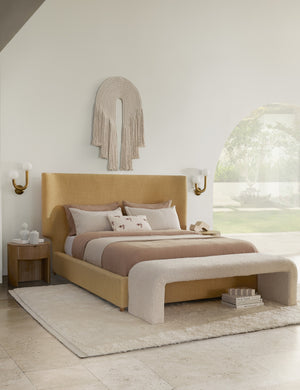 The Kono round solid oak nightstand with one drawer has white accessories on it and sits next to a gold fabric upholstered bed with tan and ivory bedding, a curved bench, and an arched woven wall hanging above the bed.